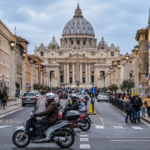 Traffico nel centro di Roma con auto e scooter in circolazione e la Basilica di San Pietro sullo sfondo.