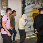 Uomo con il volto coperto accompagnato lungo una strada da alcuni carabinieri e altre persone.