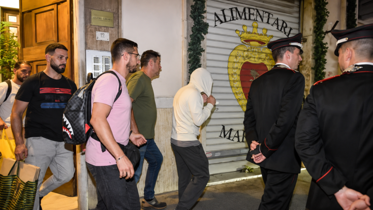 Uomo con il volto coperto accompagnato lungo una strada da alcuni carabinieri e altre persone.