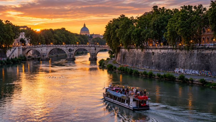 Battello naviga sul Tevere al tramonto nel centro di Roma, con un ponte storico e la cupola di San Pietro sullo sfondo.