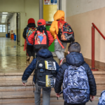 Bambini con zaini sulle spalle salgono le scale ed entrano in una scuola, accompagnati da alcuni adulti.