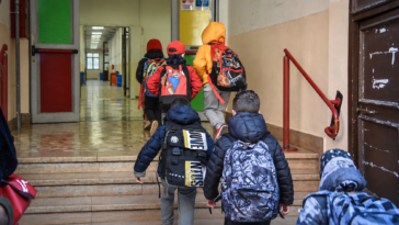 Bambini con zaini sulle spalle salgono le scale ed entrano in una scuola, accompagnati da alcuni adulti.
