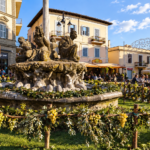 Piazza di un borgo del Lazio decorata con grappoli d’uva e rami verdi intorno a una fontana, durante una festa autunnale.