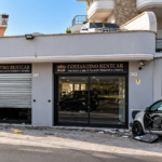Esterno di un autonoleggio con vetrata danneggiata e un’auto parcheggiata davanti al locale.