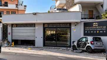 Esterno di un autonoleggio con vetrata danneggiata e un’auto parcheggiata davanti al locale.