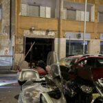 Agente delle forze dell’ordine davanti a un locale danneggiato da un’esplosione, con auto e scooter parcheggiati sulla strada.