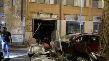 Agente delle forze dell’ordine davanti a un locale danneggiato da un’esplosione, con auto e scooter parcheggiati sulla strada.