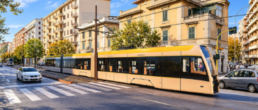 Tram moderno giallo e bianco percorre i binari lungo una strada cittadina, tra palazzi residenziali, automobili e attraversamenti pedonali.
