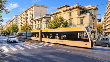 Tram moderno giallo e bianco percorre i binari lungo una strada cittadina, tra palazzi residenziali, automobili e attraversamenti pedonali.