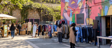 Mercatino all'aperto con stand di artigianato e abbigliamento vintage, visitatori tra gli espositori e un edificio decorato con murales colorati e insegna Teatro.